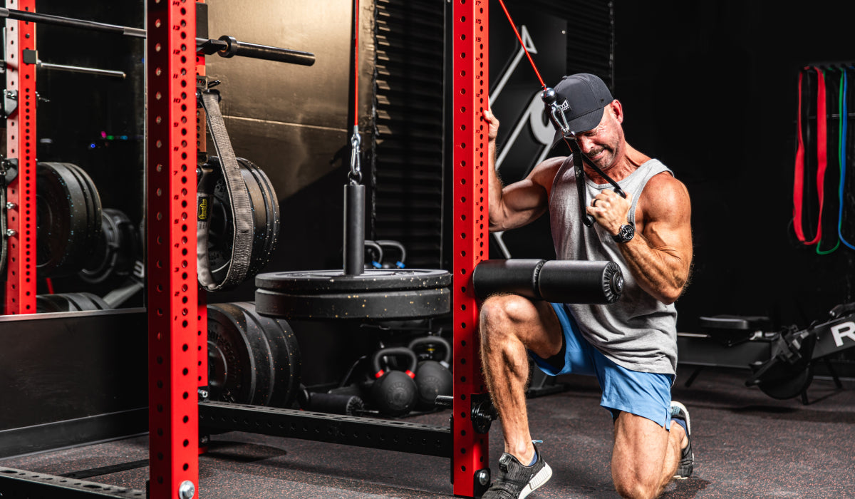 Man performing a single-arm cable crunch exercise in a gym, kneeling beside a red power rack, engaging core muscles while using a cable machine with weight plates visible in the background.