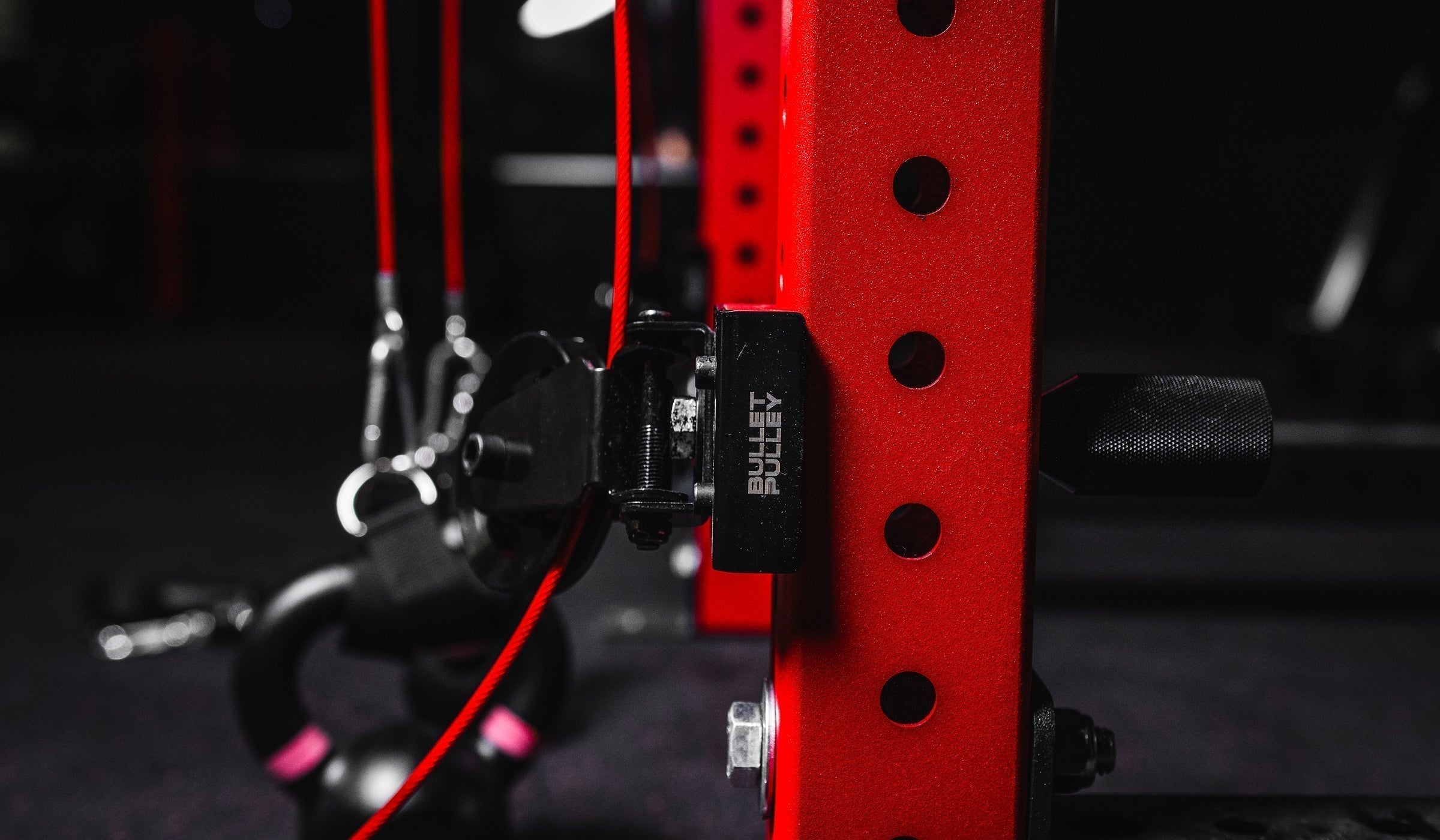 Close-up of a Bullet Pulley cable attachment mounted securely on a red power rack, with red cables and gym equipment visible in the background