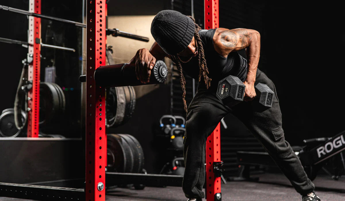 A person working out in a gym, performing a single-arm row with a dumbbell in their right hand while using a red squat rack for support. The gym features various weight equipment including kettlebells, barbells, and a rowing machine in the background.