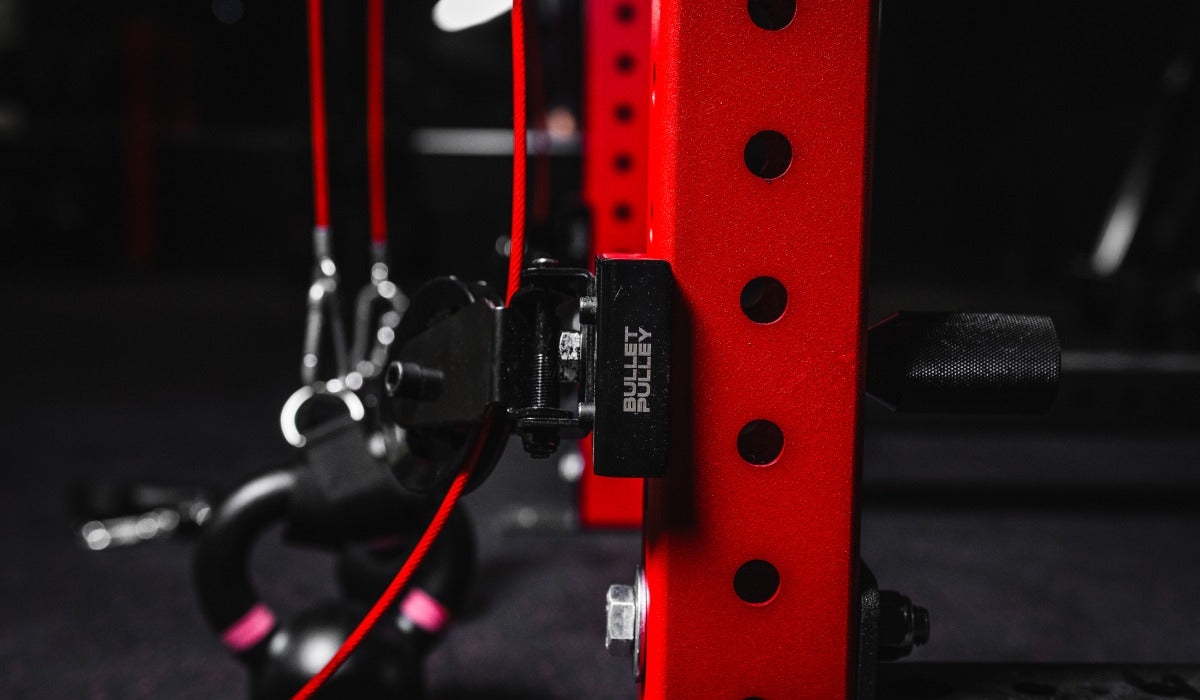 Close up of a red power rack with a mounted home gym pulley system and cable attachment in a dark gym setting.