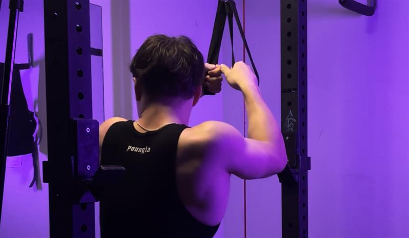 Man performing a resistance band shoulder exercise in a gym under purple lighting, wearing a black tank top with the brand name "YoungLA" on the back