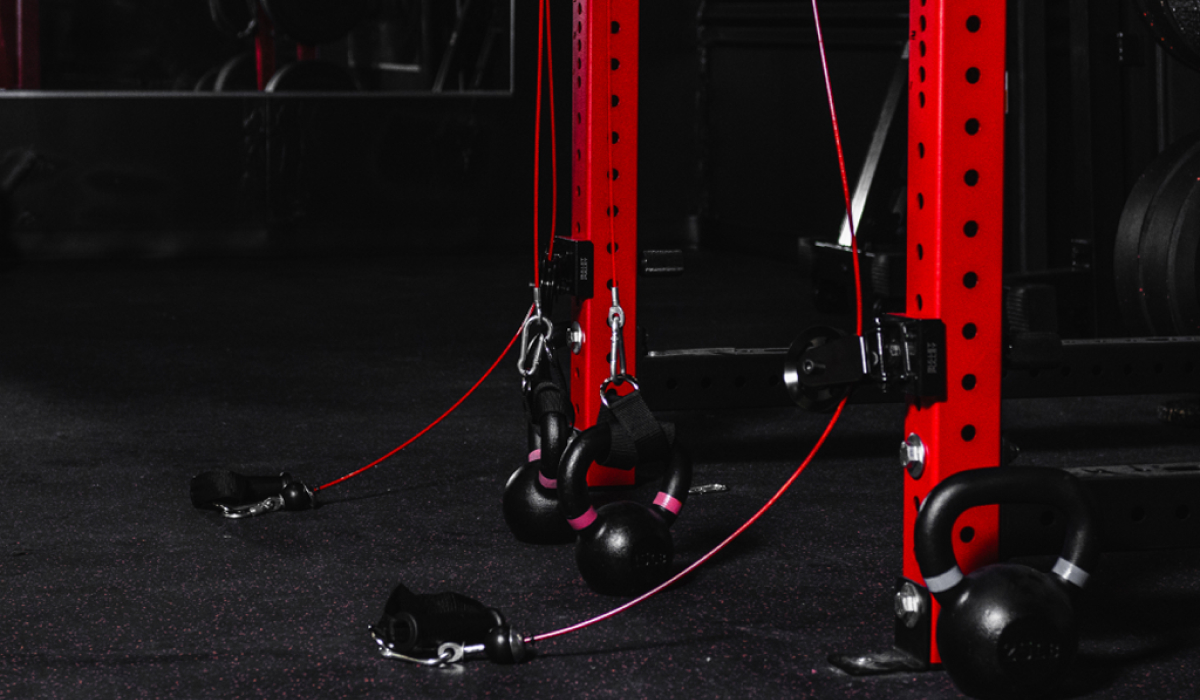Close-up of kettlebells attached to cable system in a home gym setup. Red power rack with adjustable hooks and cables for strength training exercises.