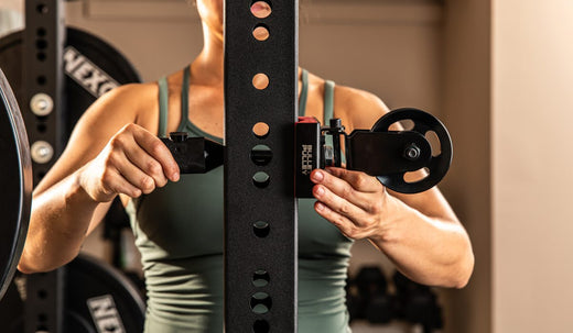 Close-up of a person adjusting equipment on a home gym power rack. The image shows the installation of a fitness accessory on a sturdy gym frame for workout customization.