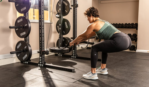 Home gym workout setup featuring a person performing a squat with a barbell. Surrounding equipment includes weight plates and a squat rack in a spacious home gym space.