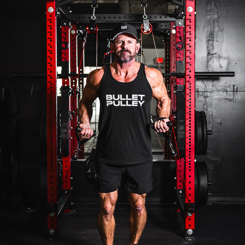 Man wearing a BULLET PULLEY tank top, performing a workout with a cable pulley system in a home gym. The home gym has a red and black color scheme, with weights and pulleys visible in the background.