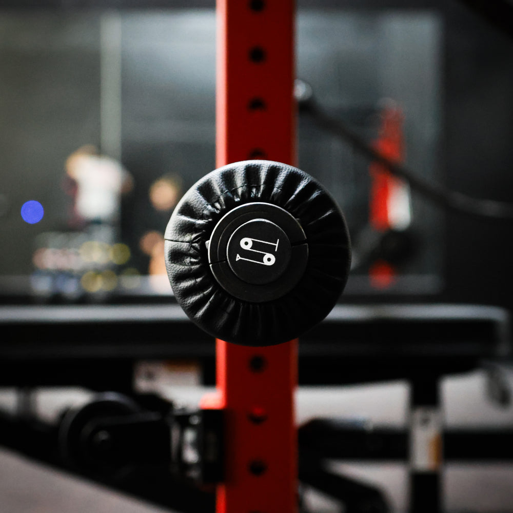 Close-up of a gym equipment's padded barbell holder, featuring a black foam cushion with a logo on a red rack. The background is blurred, showcasing a well-lit home gym environment.