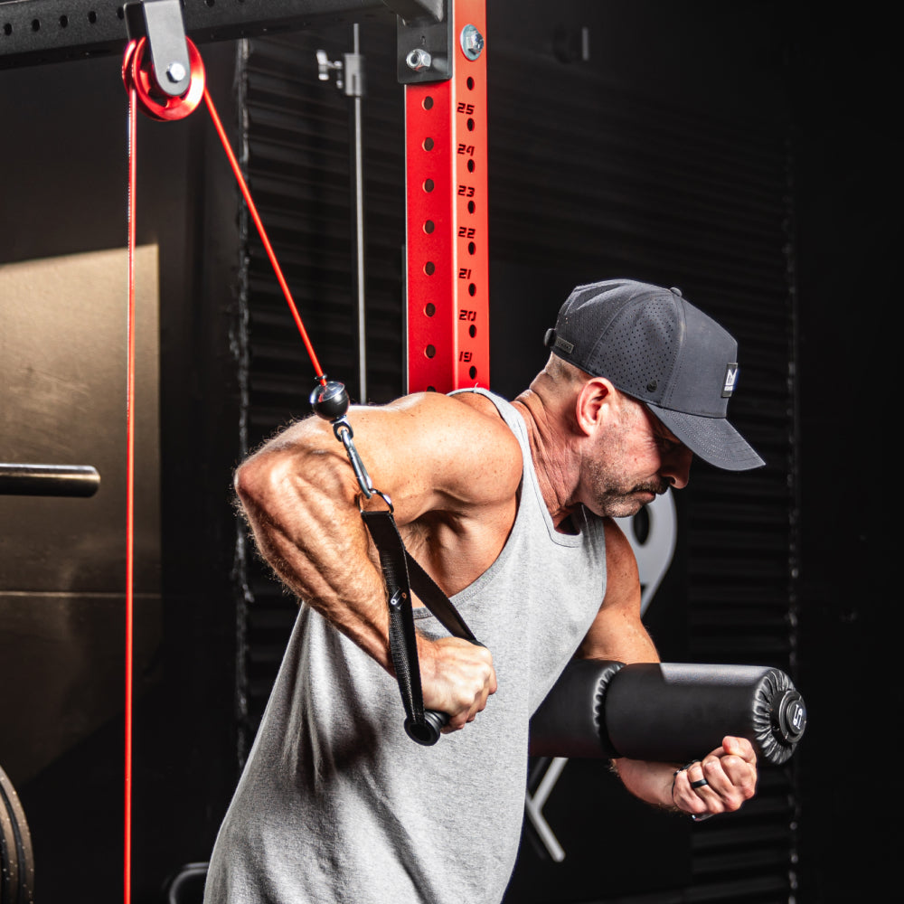 Man performing triceps pushdowns with cable machine at home, wearing a gray tank top and black cap, focusing on strength training exercise.