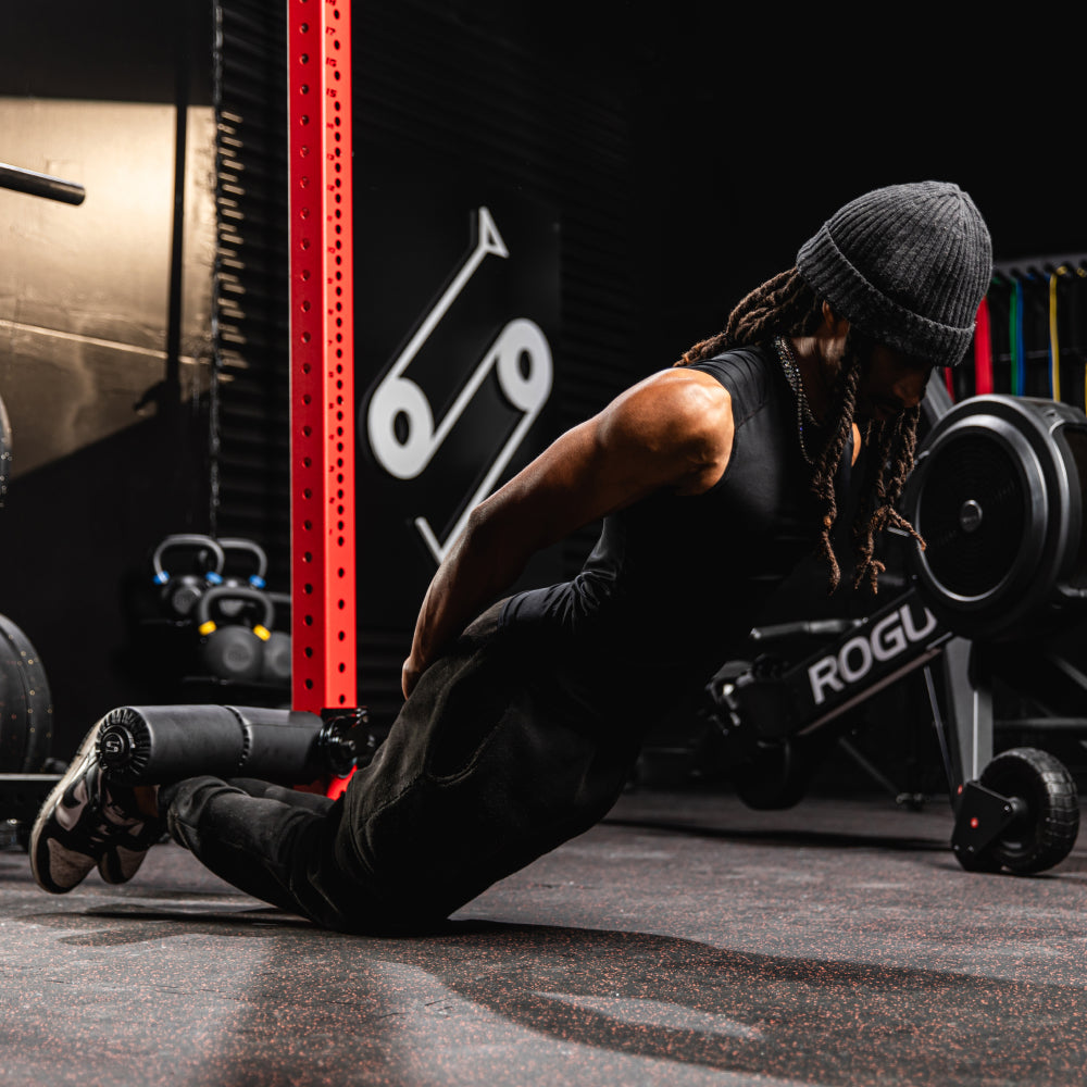 Person performing a tricep dip exercise in a gym, wearing a beanie, black sleeveless top, and black pants. Home gym equipment like kettlebells and a rowing machine (Rogue brand) are visible in the background. The gym has a modern, dark aesthetic with rubber flooring.