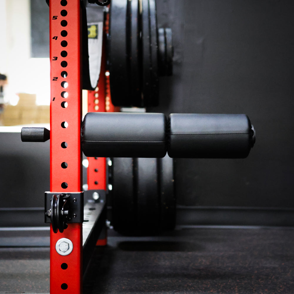 Close-up image of a squat rack with black foam padding and weight plates in the background, showcasing the home gym equipment's red frame and adjustable features for leg exercises.