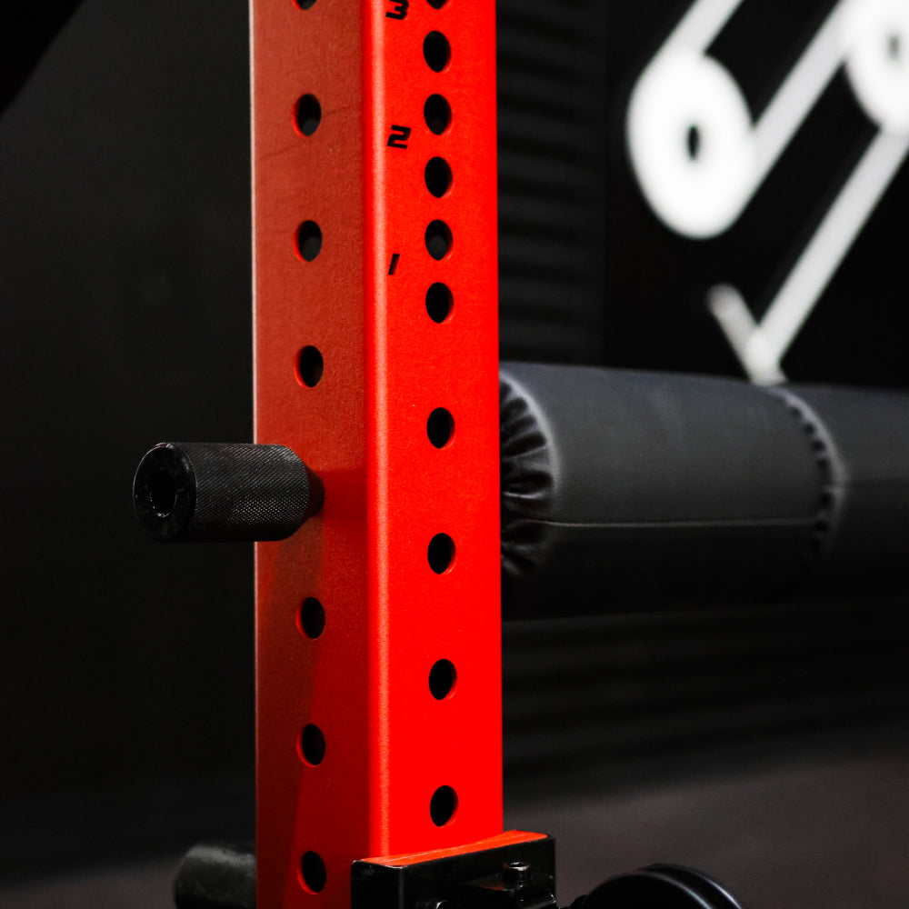 Close-up of a red gym equipment frame with numbered hole adjustments for barbell placement, featuring black padding on the horizontal bars for support, set against a dark background.