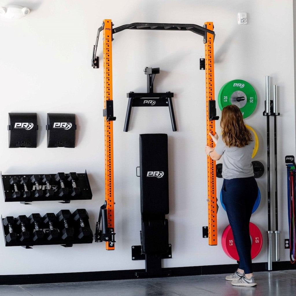 Home gym setup featuring a PRX Performance folding rack, dumbbell rack, weight plates, and resistance bands. A woman adjusts the height of the rack while surrounded by various fitness equipment, including dumbbells and a bench.