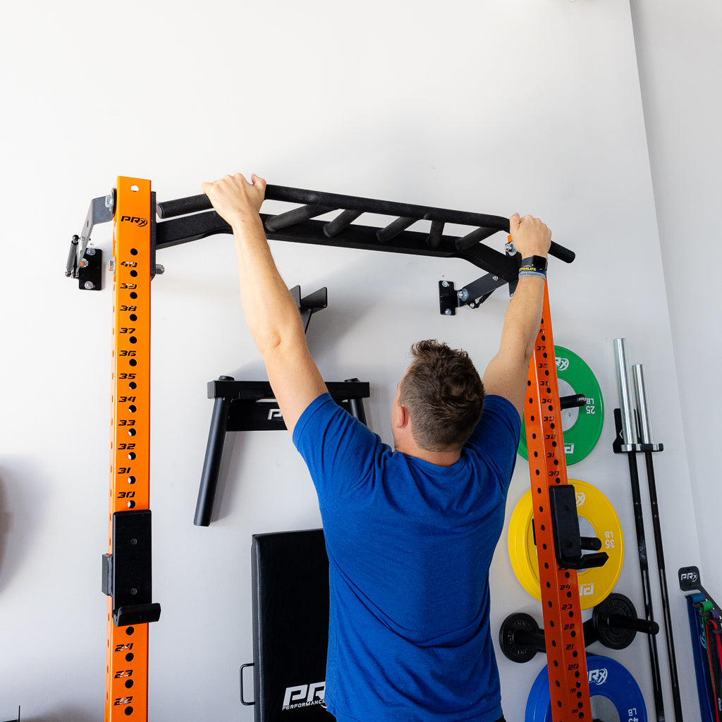 Person performing a pull-up exercise on a squat rack with a pull-up bar in a home gym setting. The orange rack is marked with measurement markings, and weight plates in various colors are visible in the background.