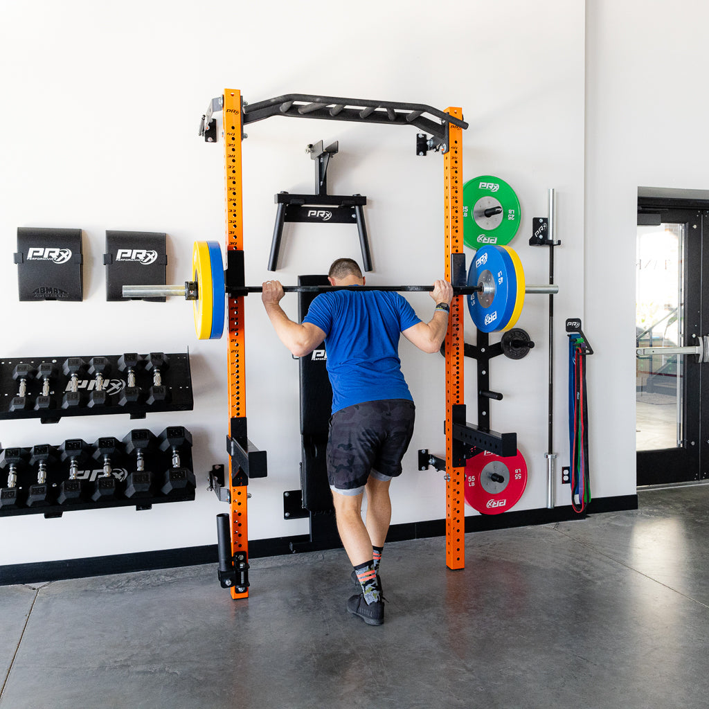 A person performing a squat exercise with a barbell in a home gym, using a Pro squat rack with a pull-up bar. The home gym setup includes dumbbells, weight plates in various colors, resistance bands, and a black padded support for the barbell. The home gym has a clean, modern look with concrete floors and bright orange squat rack posts.