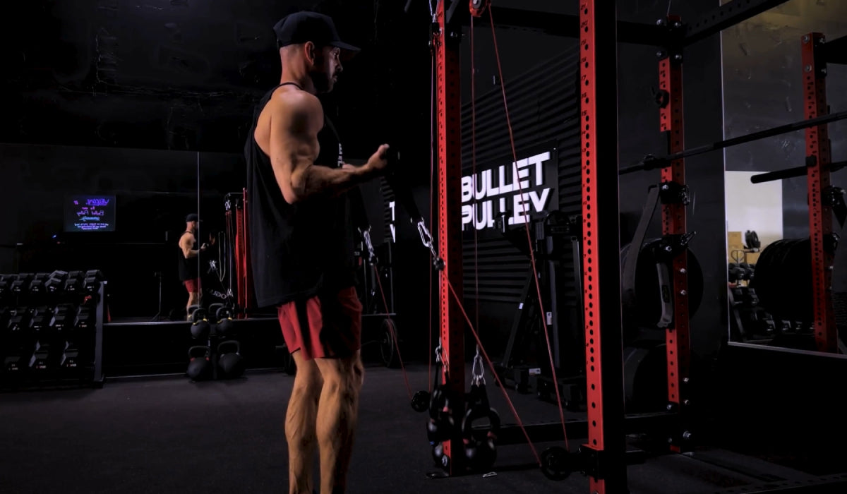 A man training in a home gym using a Bullet Pulley cable system attached to a red power rack, performing an arm exercise in a focused indoor workout space.
